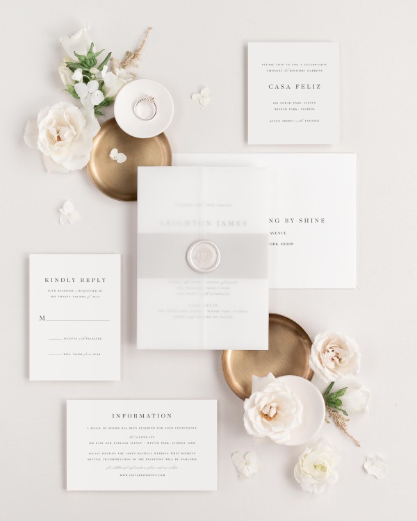 Leighton wedding invitations flat lay with feather belly band, translucent vellum jacket, and pearl florette wax seal.