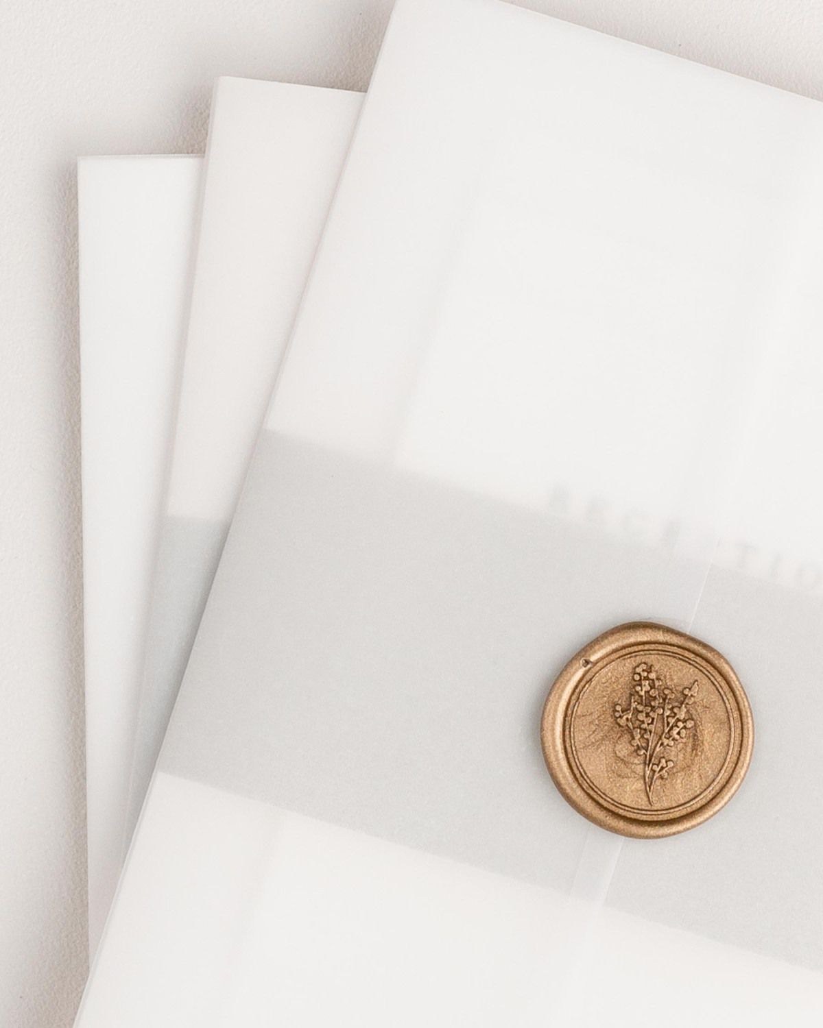 A small pile of Poppy wedding invitation suites assembled with stone belly bands, wrapped with vellum jackets, and secured with a gold posy wax seal.