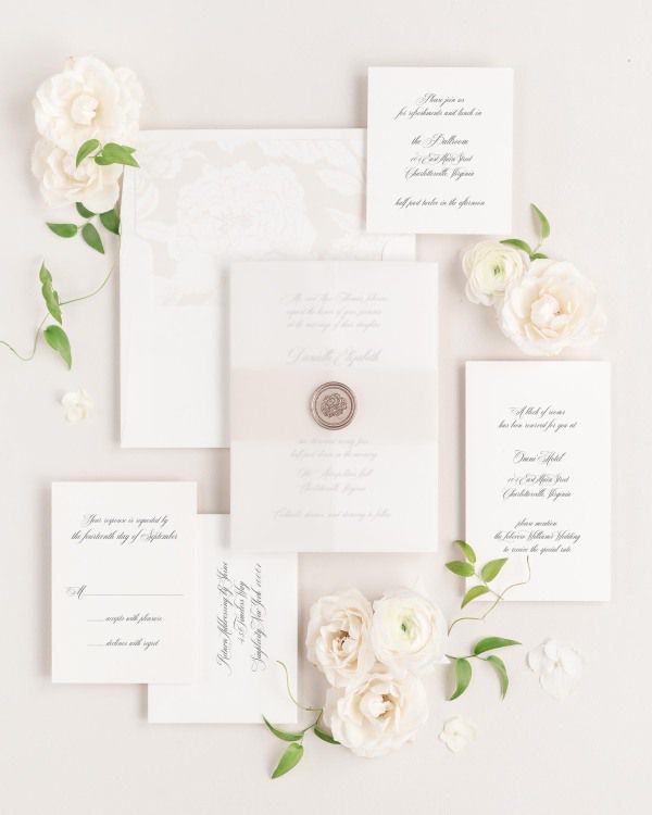 A Danielle wedding invitation suite is beautifully placed on a neutral background with flowers and greenery. The wedding invitation is wrapped with a belly band and vellum jacket, and is sealed with a bronze florette wax seal.